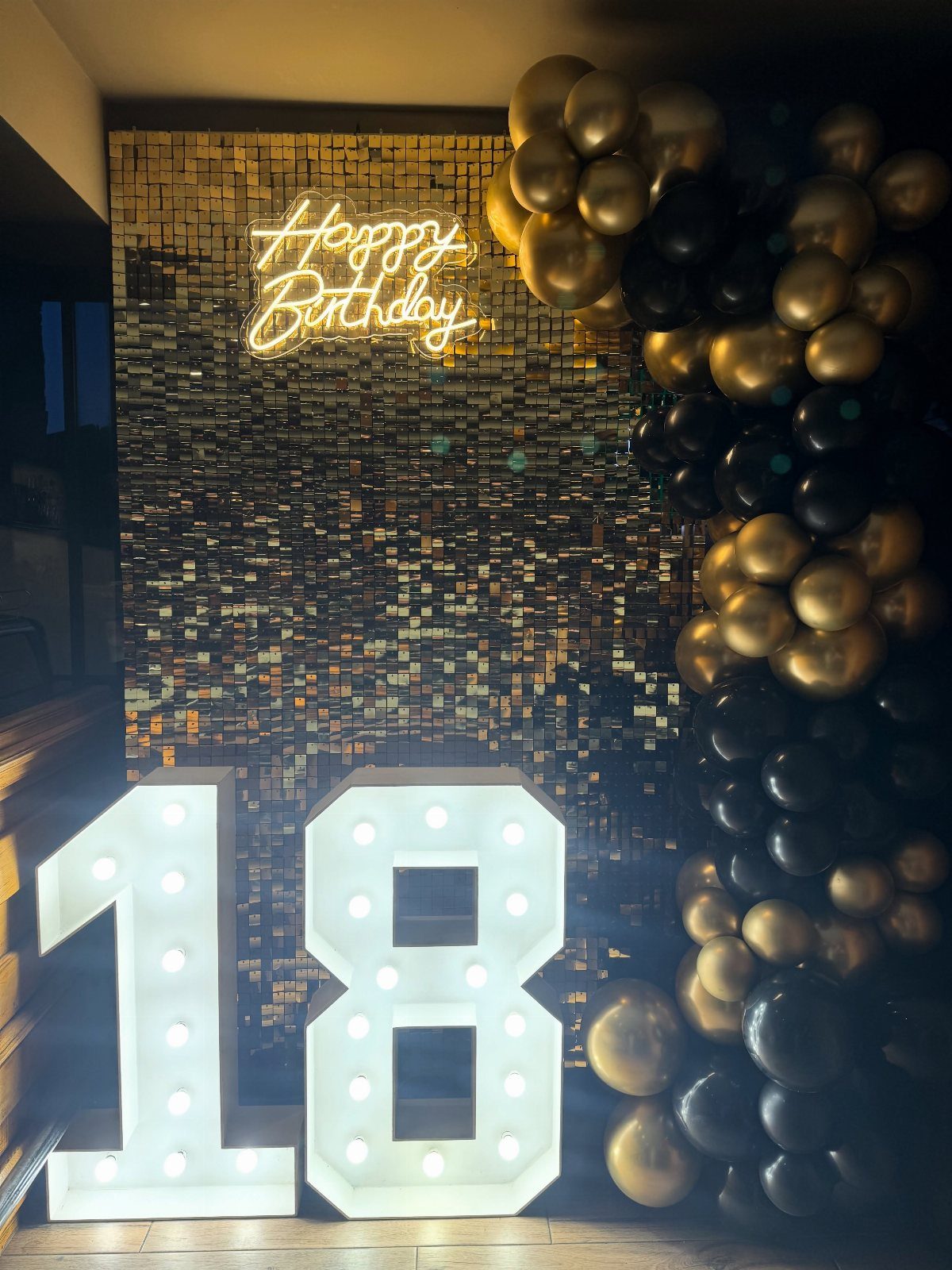 Illuminated number 18 with gold sequin backdrop and black and gold balloons.