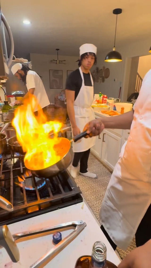 Young men in chef attire cooking with a large flame in a kitchen