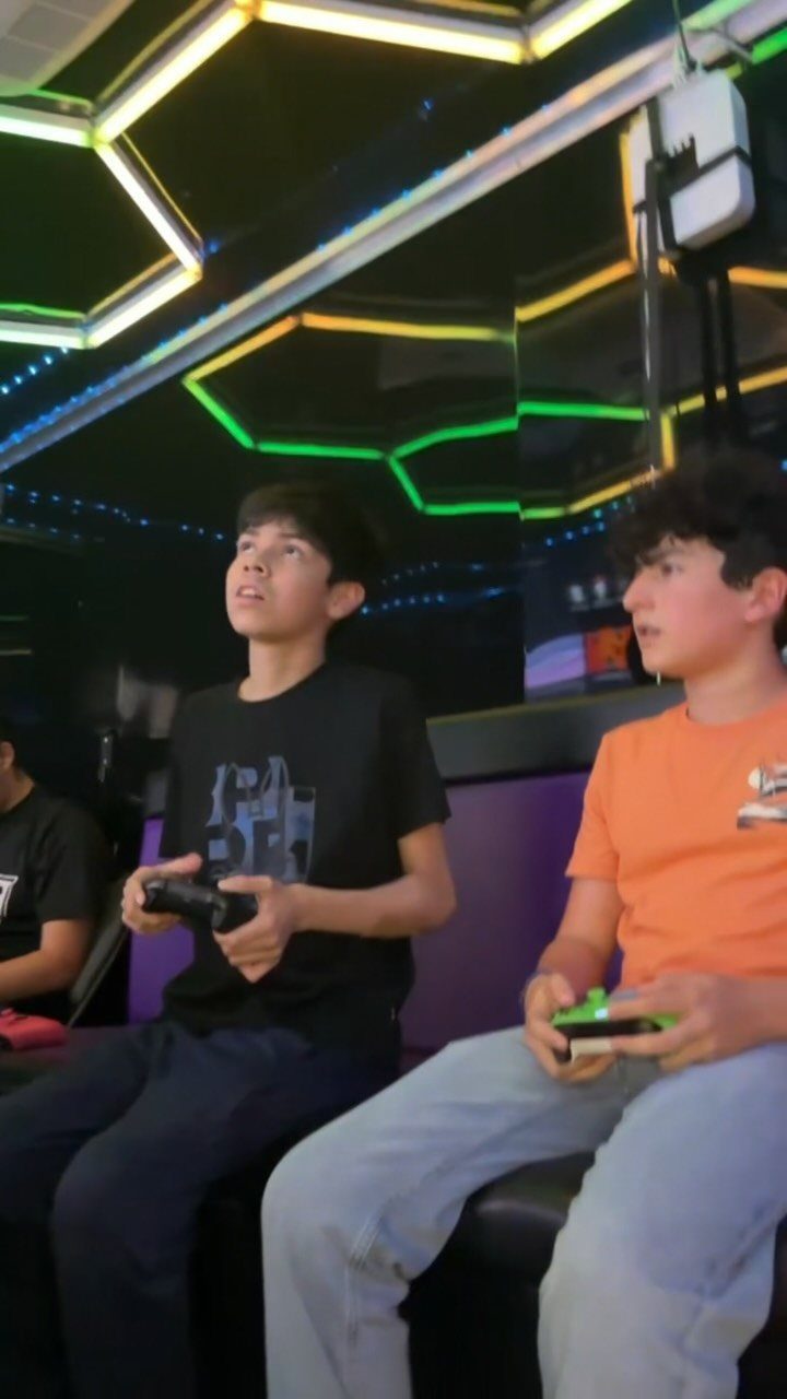 Two teenage boys playing video games in a neon-lit gaming lounge.
