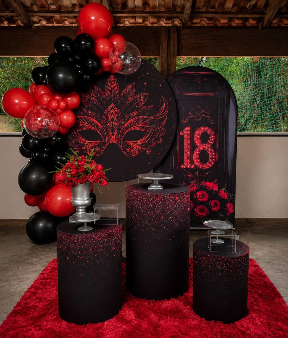 Birthday party decoration with red and black balloons, pedestals, and a masquerade mask backdrop.