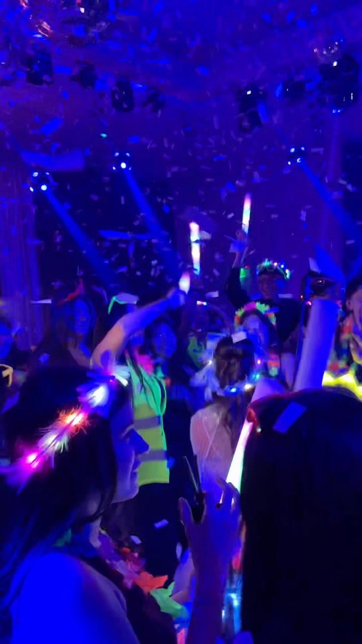Young people dancing with neon glow sticks and headbands under blue party lights.