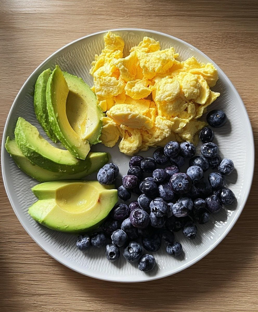 Scrambled eggs, sliced avocado, and blueberries served on a white plate on wood.