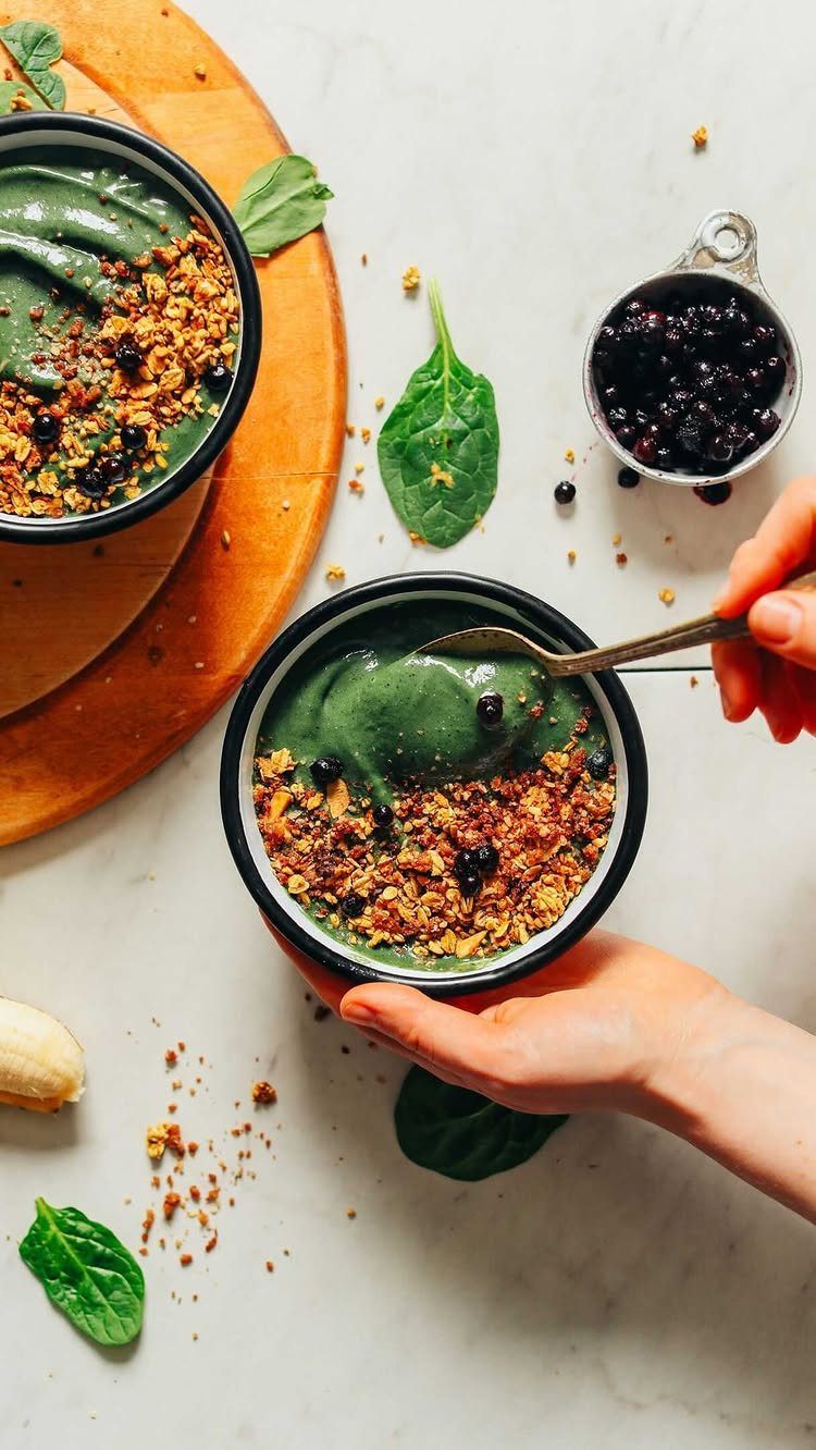 Green smoothie bowls topped with granola and blueberries on a white marble surface.