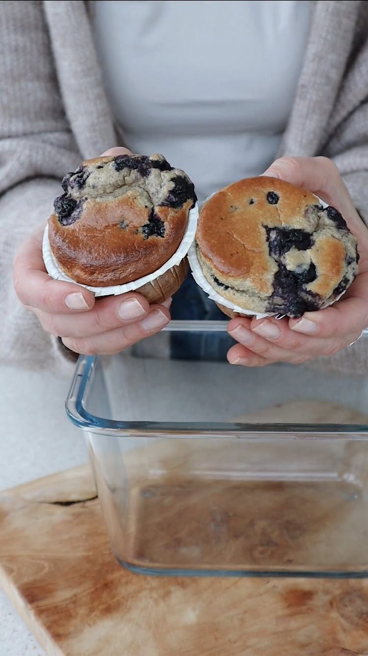 Two blueberry muffins held in hands over a clear glass baking dish.