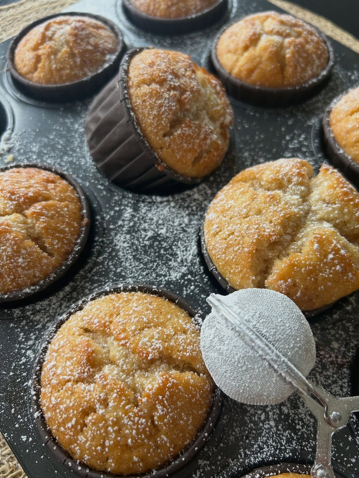 Golden brown muffins in a baking tin dusted with powdered sugar and a sifter.