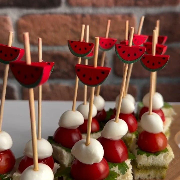 31 Beach Themed Appetizers for Parties, Picnics and Pool Days 16 Caprese skewers with watermelon-shaped picks arranged against a red brick wall background.