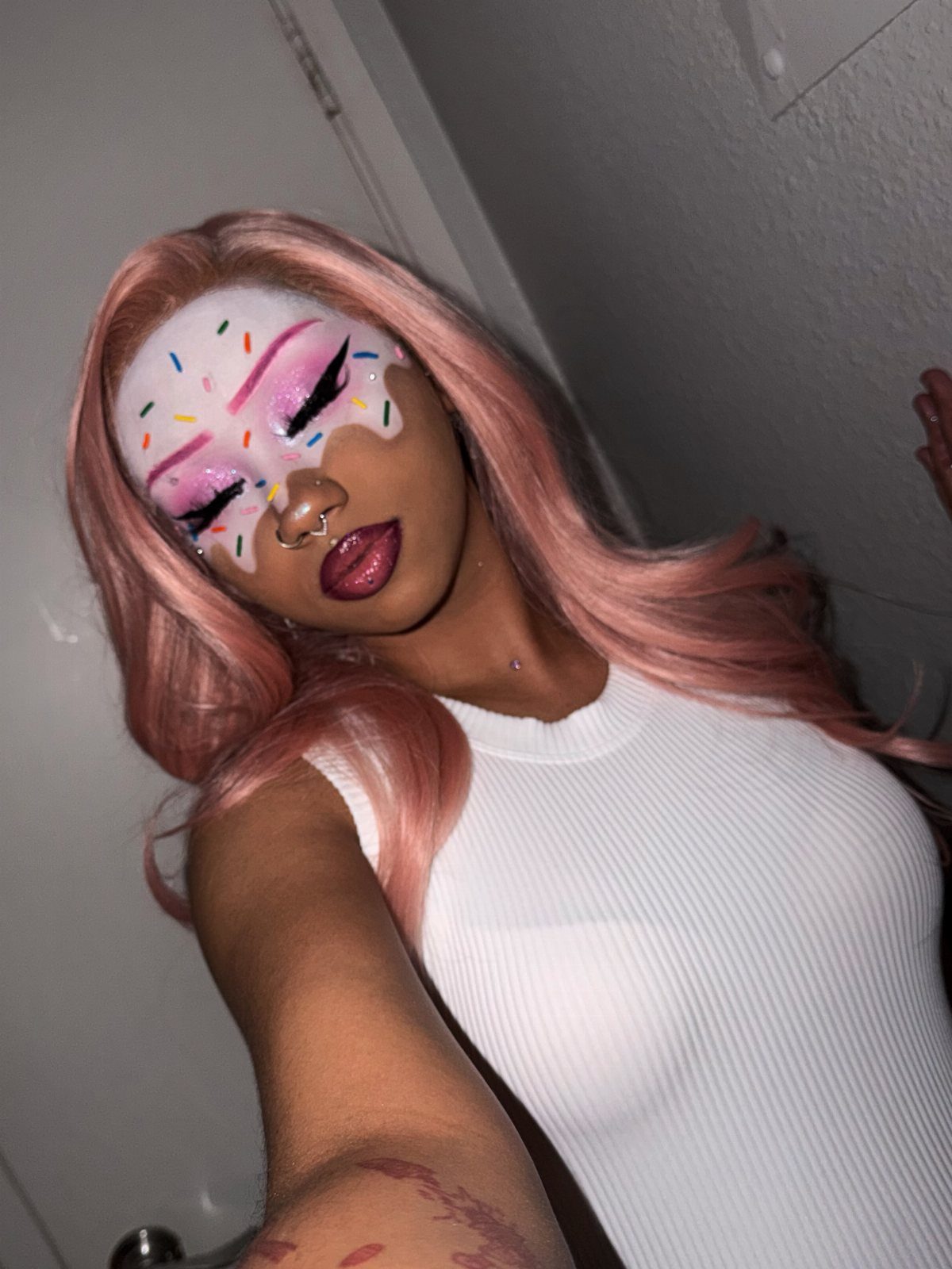 Person with pink hair wearing white frosting face paint with colorful sprinkles.