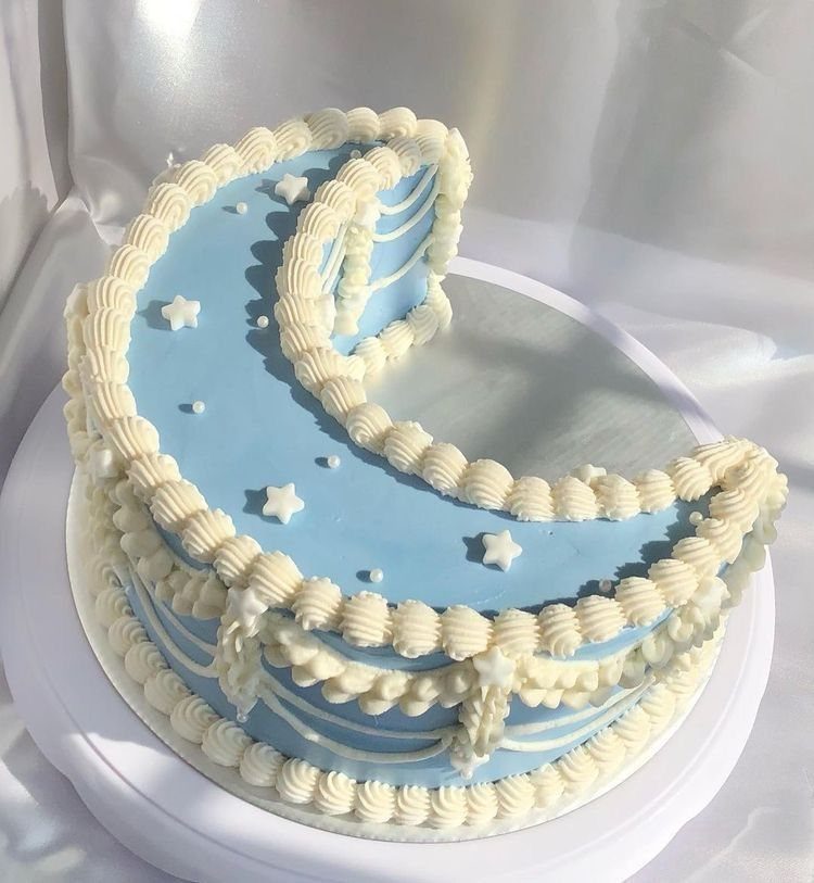 Crescent moon-shaped light blue cake with white cream piping and small star sprinkles.