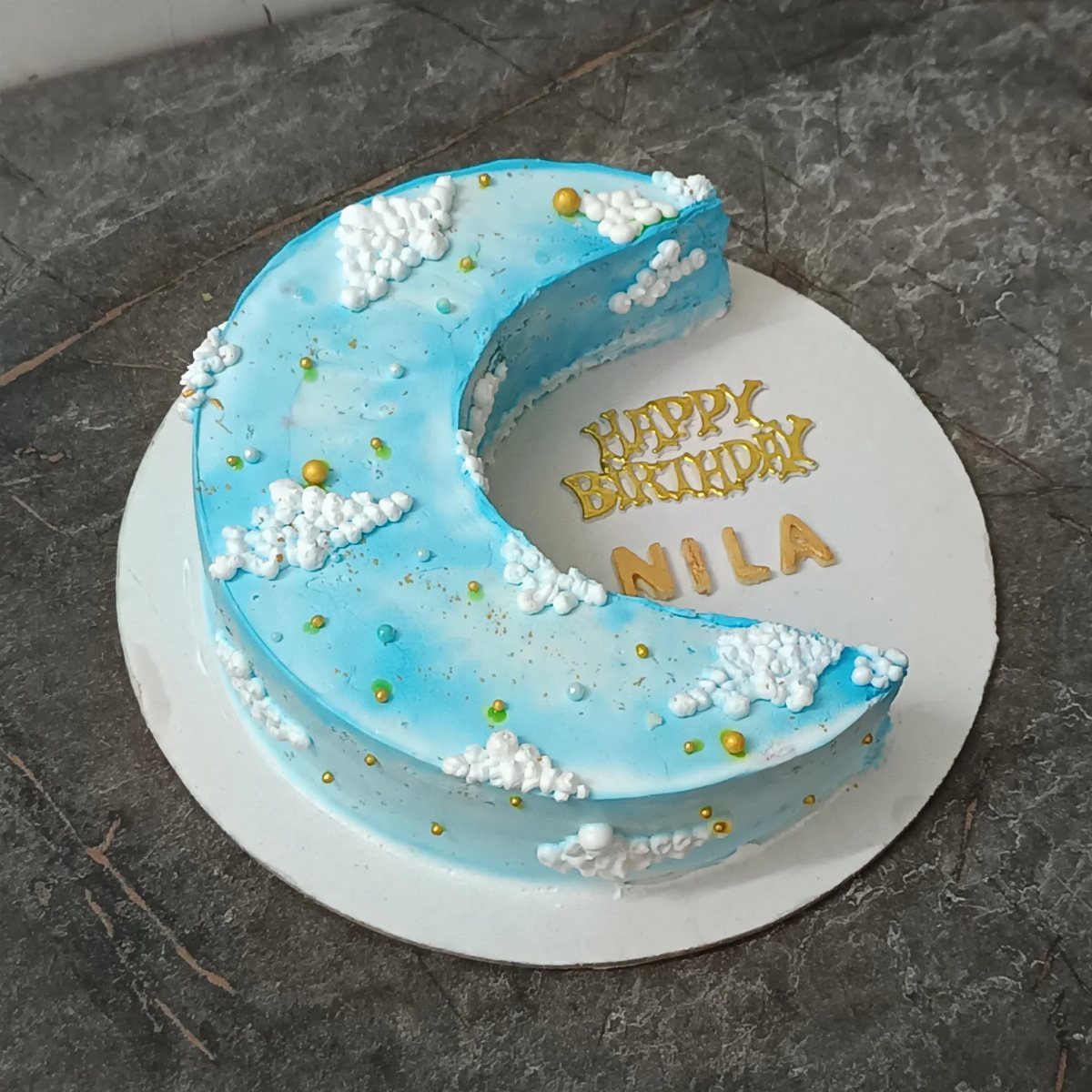 Crescent moon-shaped blue cake decorated with white frosting clouds and gold pearls.