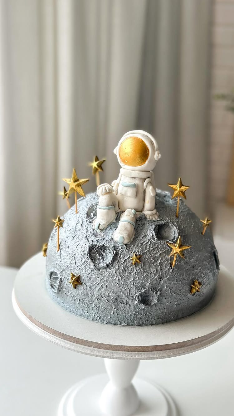 Grey moon-textured cake with a small astronaut figure and gold stars on a stand.