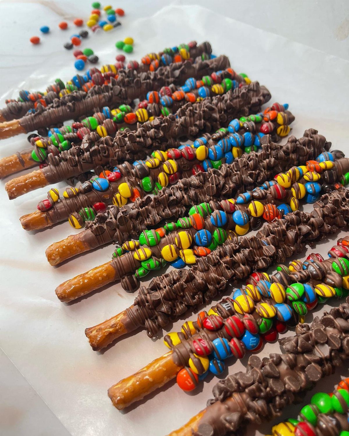 13 Birthday Party Treat Ideas, From Popcorn to Tea Sandwiches 2 Chocolate-dipped pretzel rods decorated with colorful candy-coated chocolate pieces on white parchment paper.