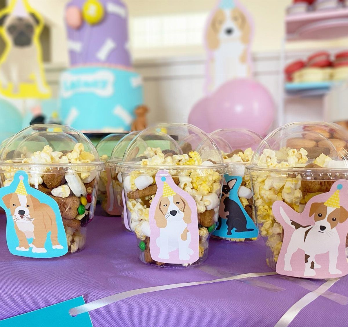 13 Birthday Party Treat Ideas, From Popcorn to Tea Sandwiches 6 Puppy chow snack mix in clear plastic cups with dog stickers on purple tablecloth