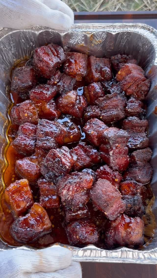 25 Black People Finger Foods 19 Glazed reddish-brown meat cubes in a silver foil tray held by gloved hands.