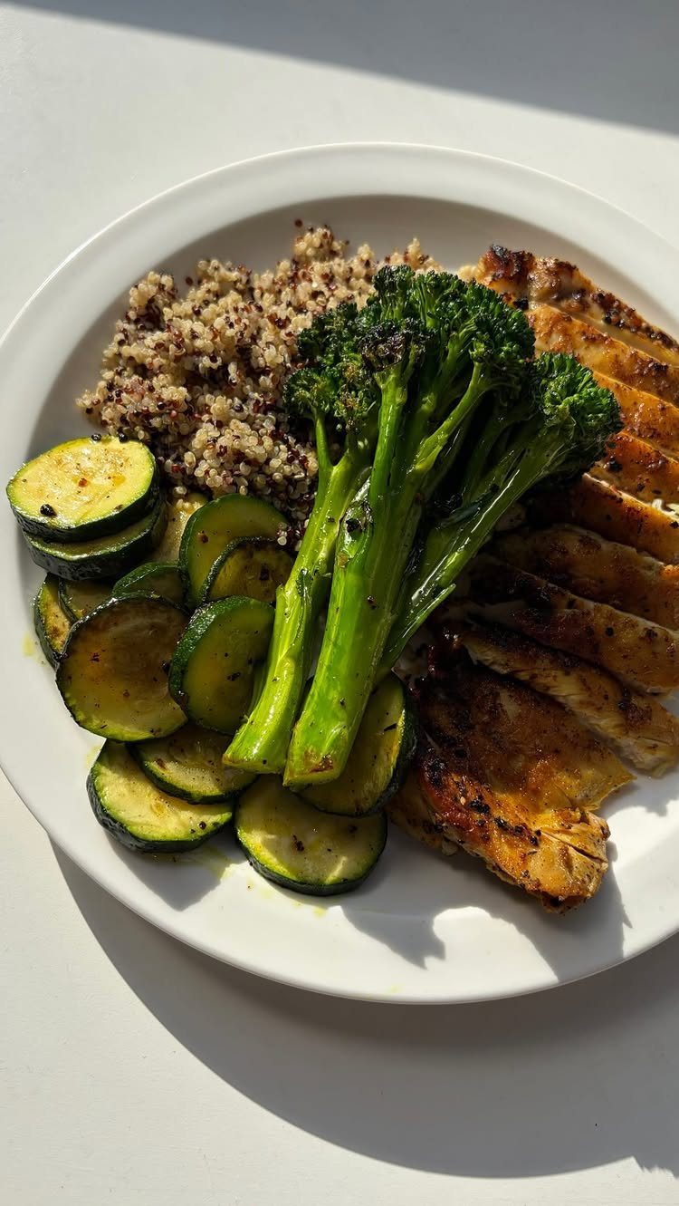 Grilled chicken breast served with quinoa, broccoli, and sliced zucchini on a white plate.