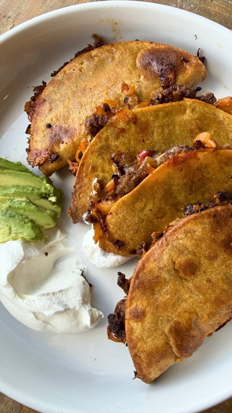 Four fried beef tacos with avocado and sour cream on a white plate
