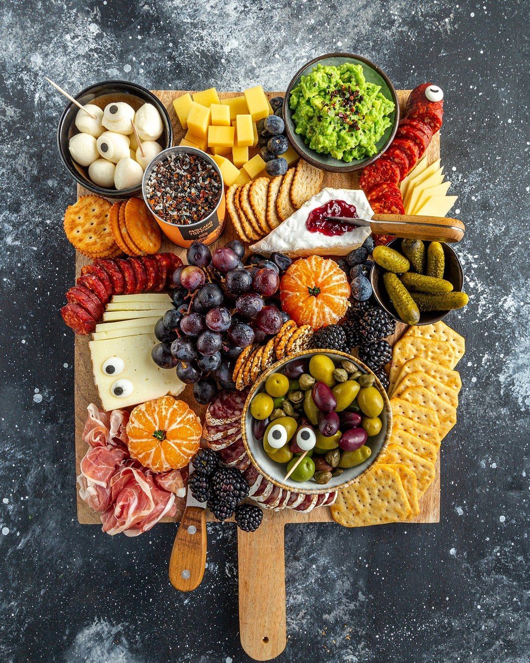 Charcuterie board with cheeses, meats, fruits, and a small bowl of cornichons.