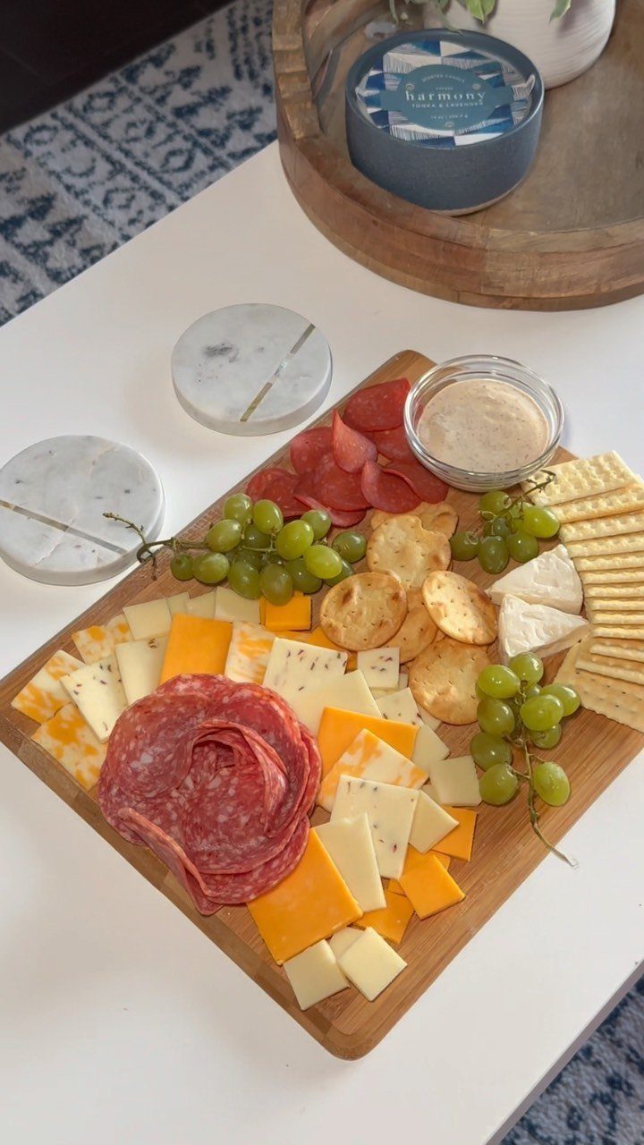 Wooden charcuterie board with salami, various cheeses, green grapes, crackers, and creamy dip.