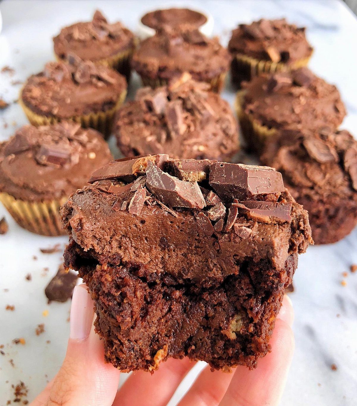 Bitten chocolate cupcake with frosting and chocolate chunks held in a hand.
