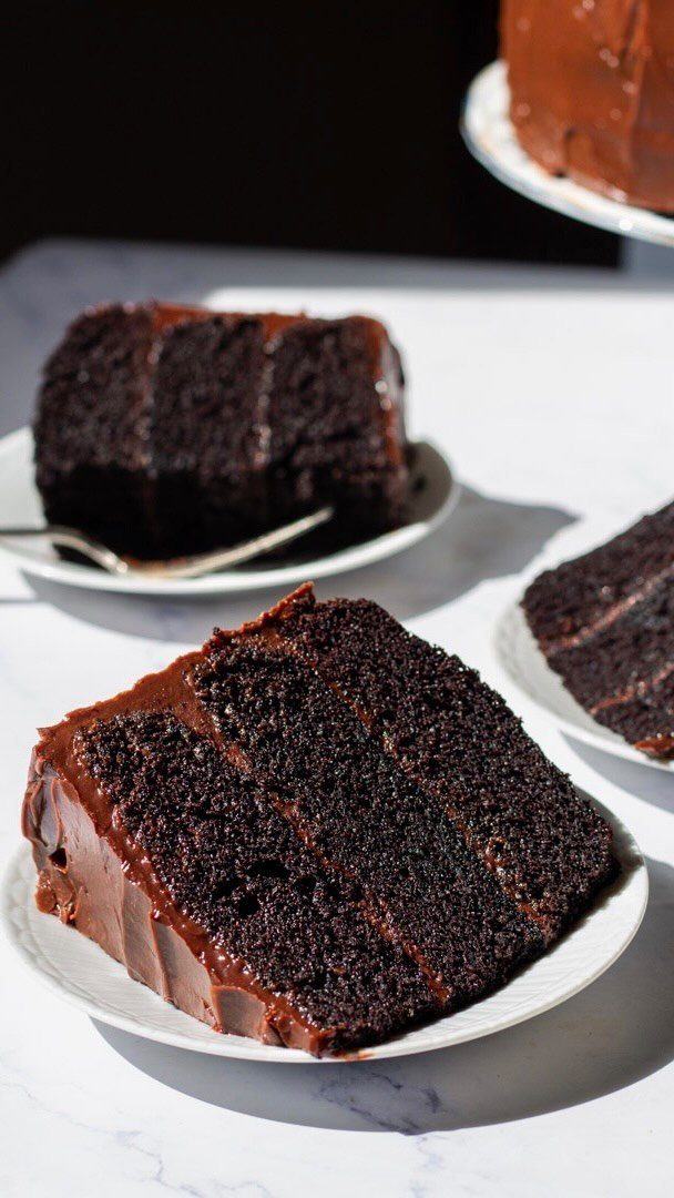 Three-layer dark chocolate cake slice with rich frosting on a white plate.