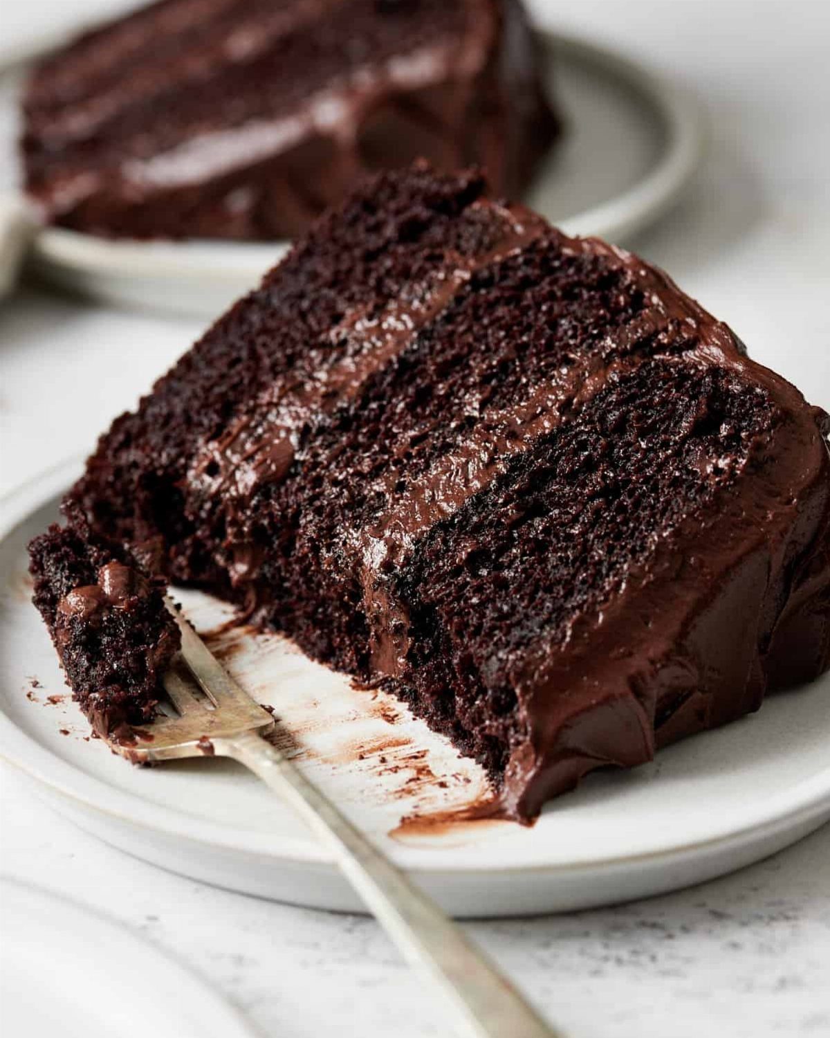 Three-layer chocolate cake slice with rich frosting on a white ceramic plate.