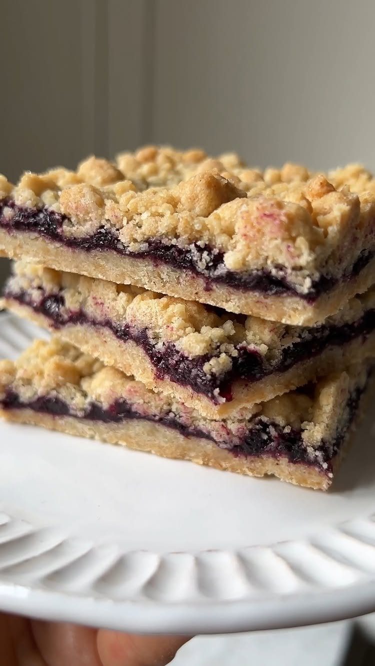 24 Desserts to Make with Blueberries, Cobbler to Cheesecake 3 Stacked blueberry dessert bars with crumbly topping on a white scalloped plate.