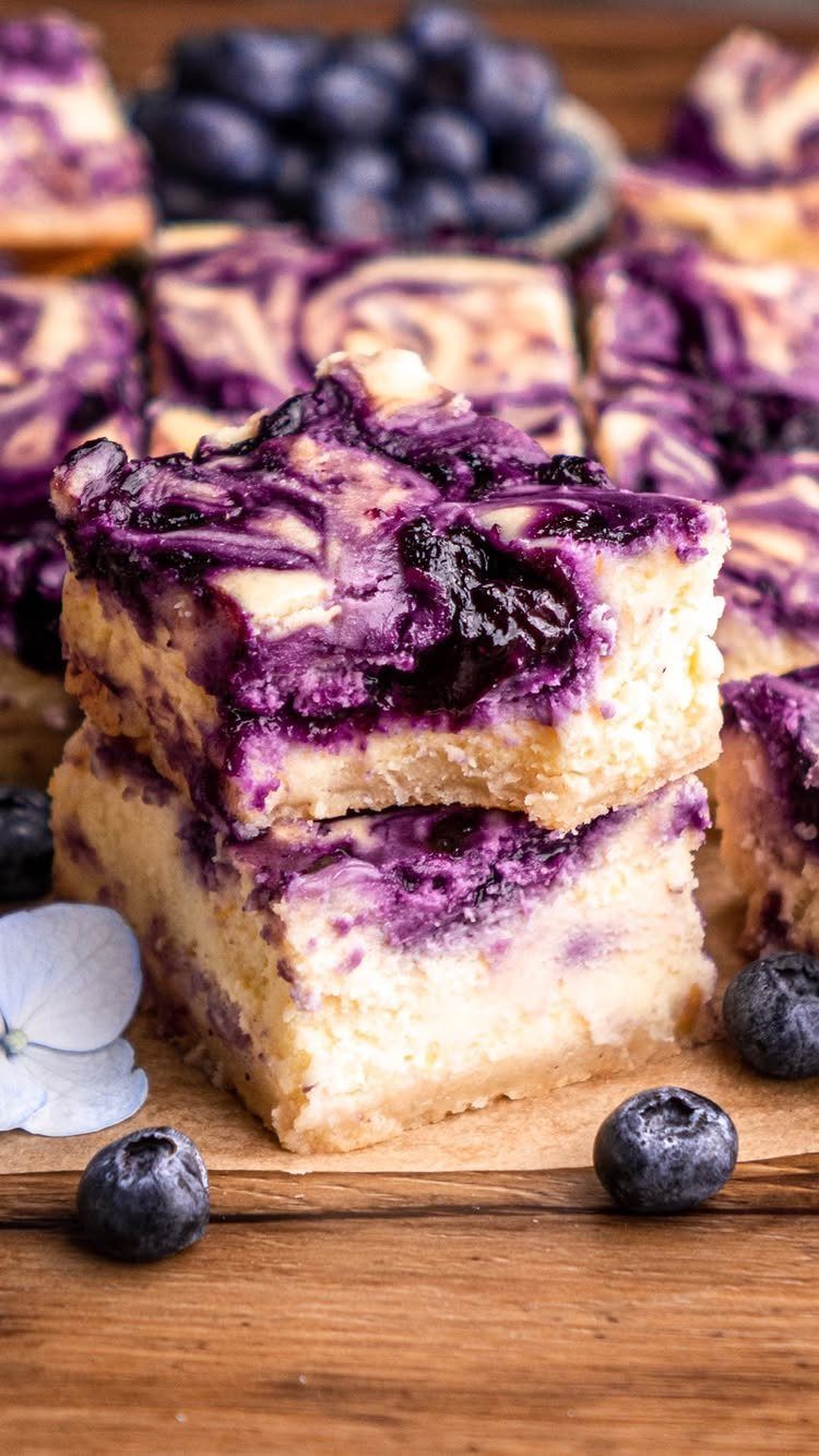 24 Desserts to Make with Blueberries, Cobbler to Cheesecake 20 Stacked blueberry cheesecake bars with purple swirls on a rustic wooden background.