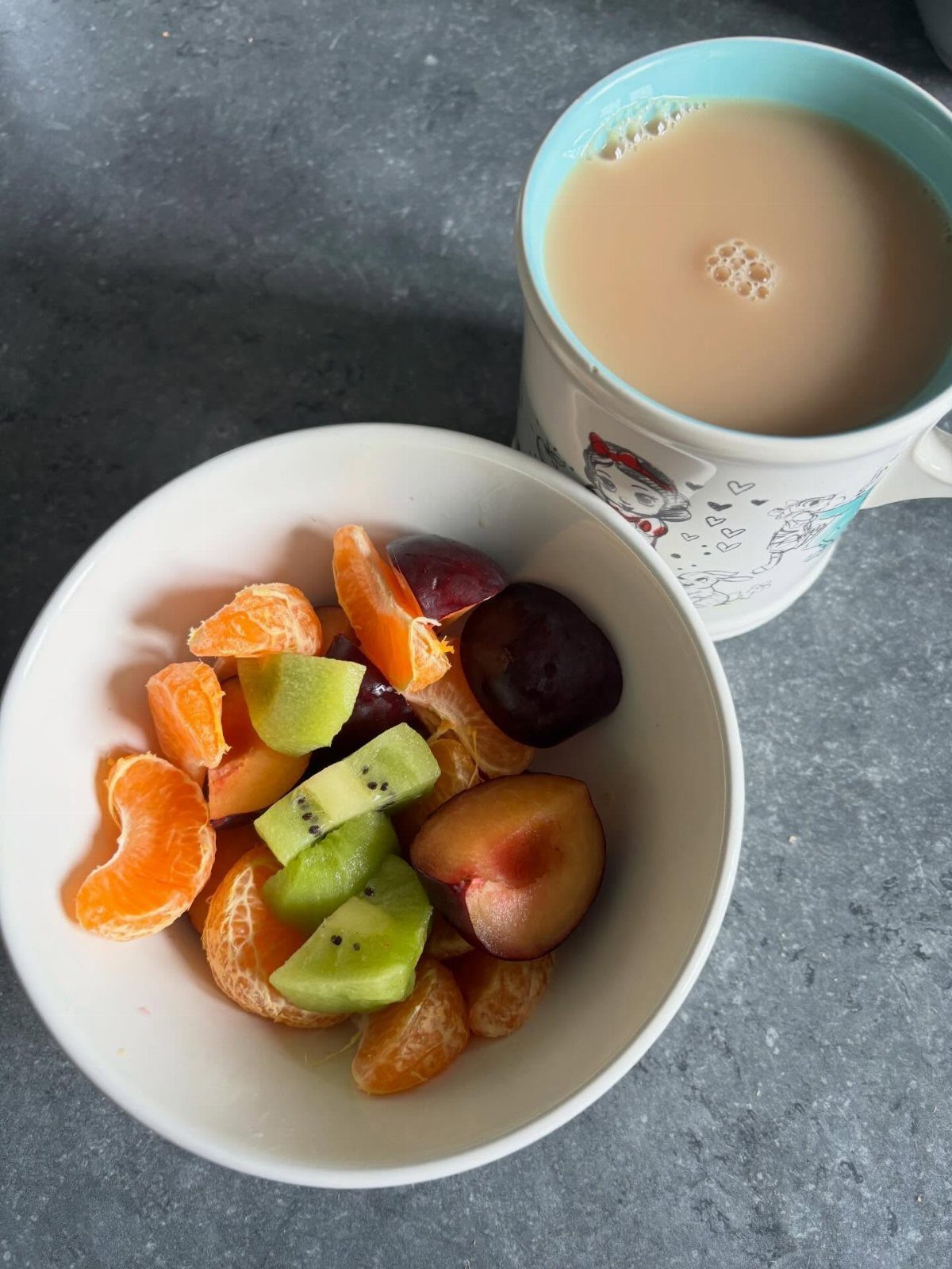 Bowl of sliced kiwi, mandarin oranges, and grapes next to a mug of tea.
