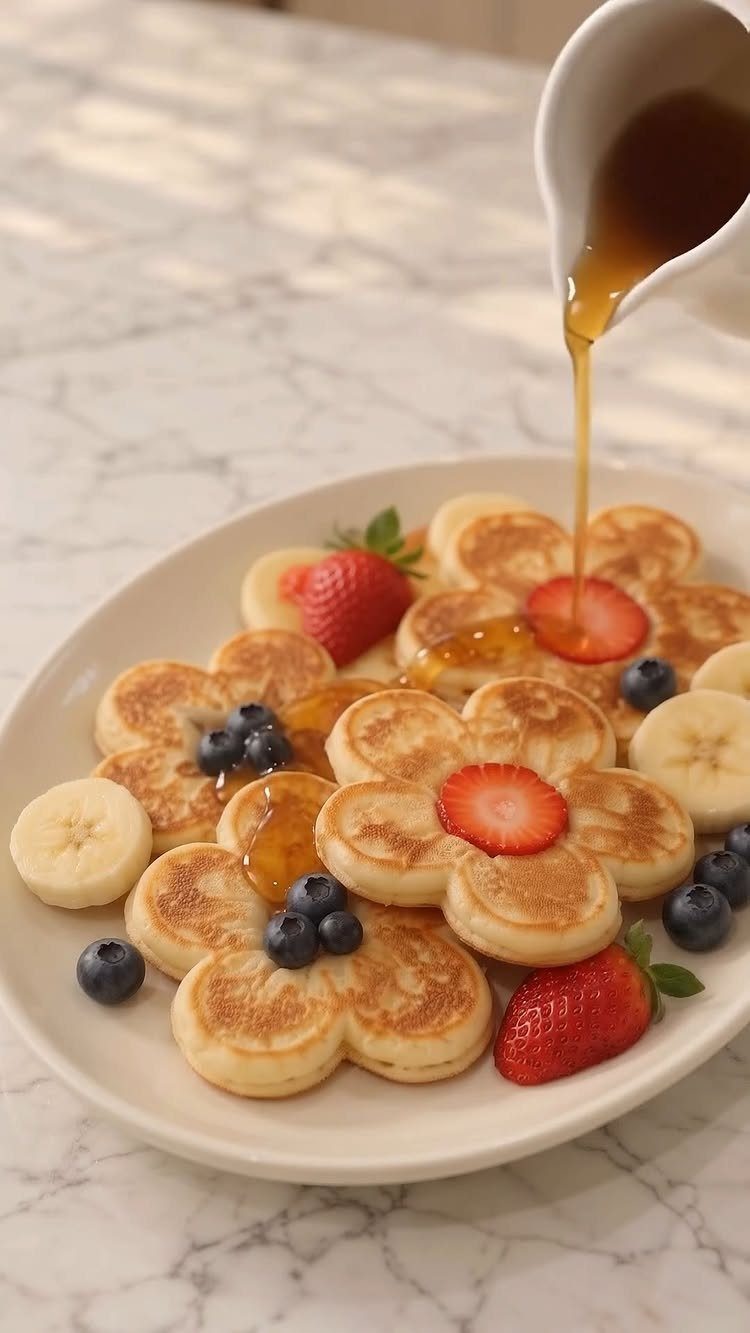 Flower-shaped pancakes topped with strawberries, blueberries, and bananas with maple syrup being poured.