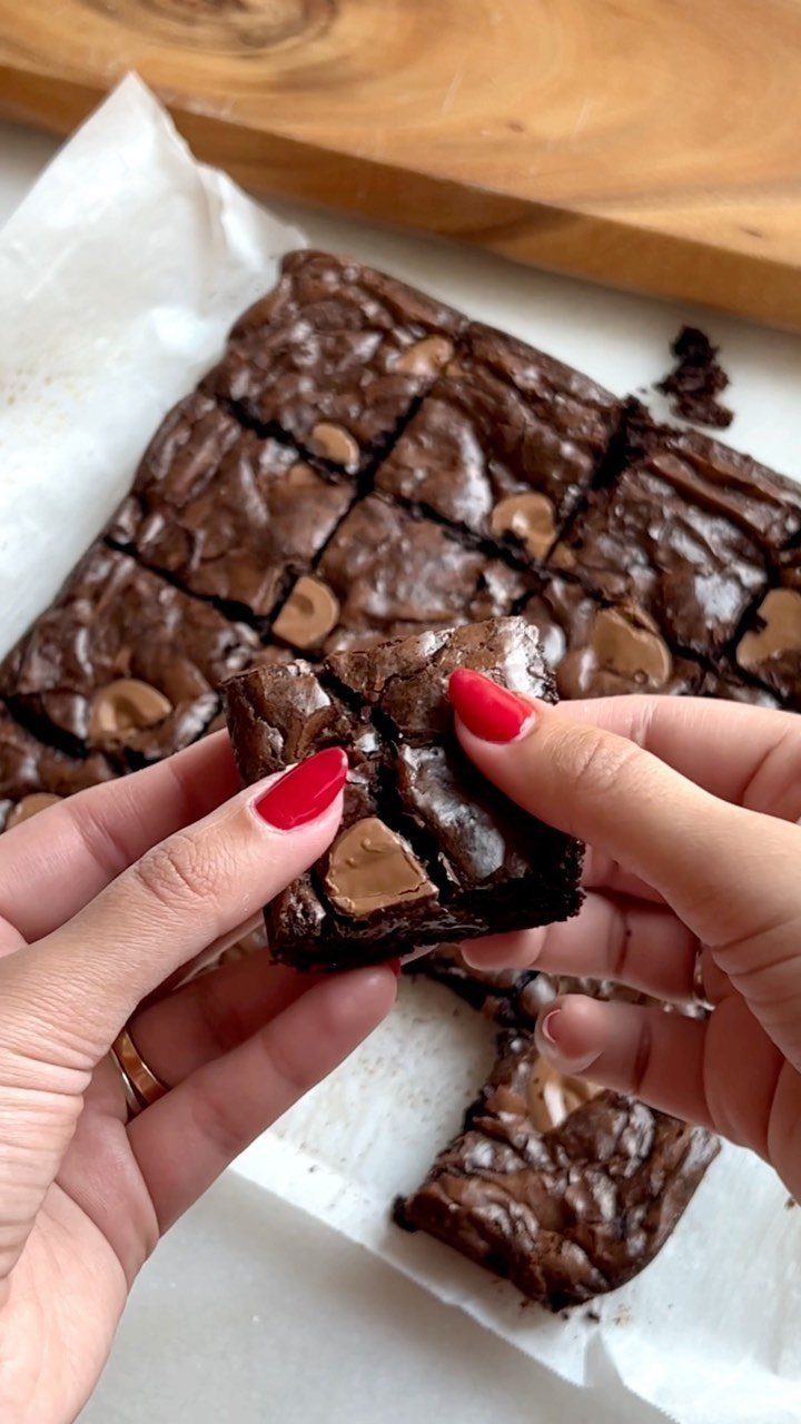 Fudgy chocolate brownies with chocolate chunks held by hands with red nails.