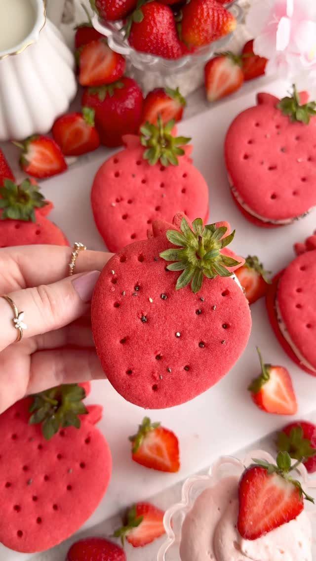 Strawberry-shaped red cookies with green leaves and fresh strawberries on a white surface.