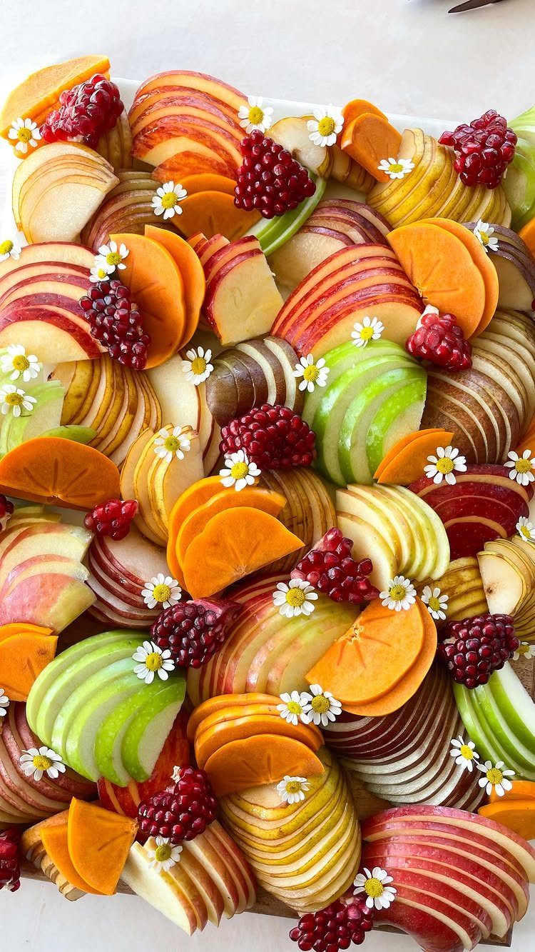 Fruit tray with sliced apples, persimmons, pomegranate seeds, and small white flowers.