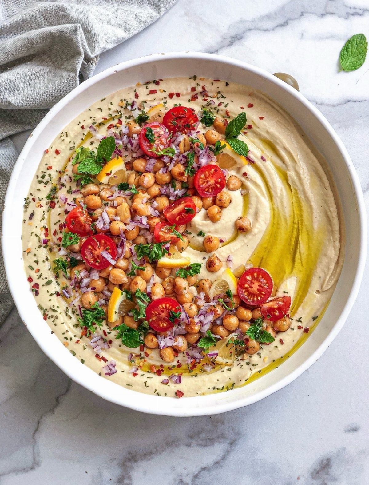 Hummus in a white bowl topped with chickpeas, cherry tomatoes, and olive oil.