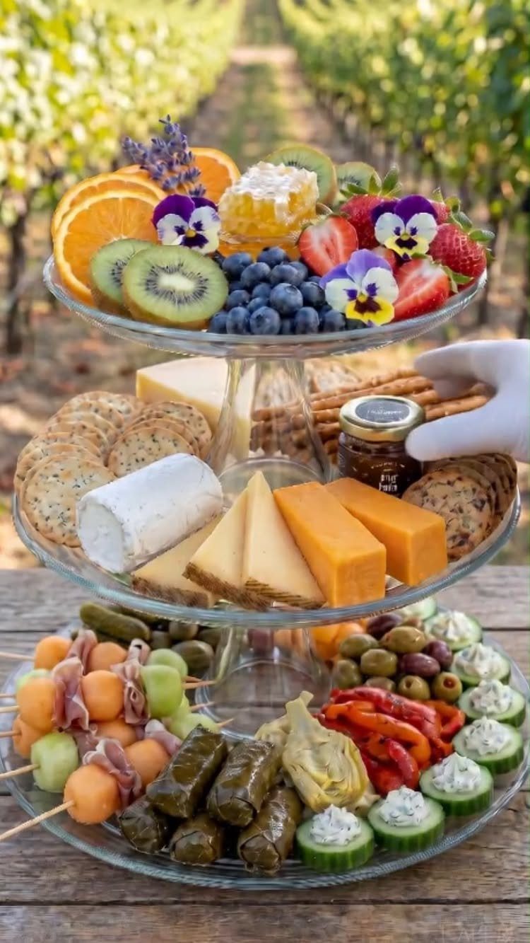 Three-tiered glass platter with fruit, cheeses, crackers, olives, and stuffed grape leaves.