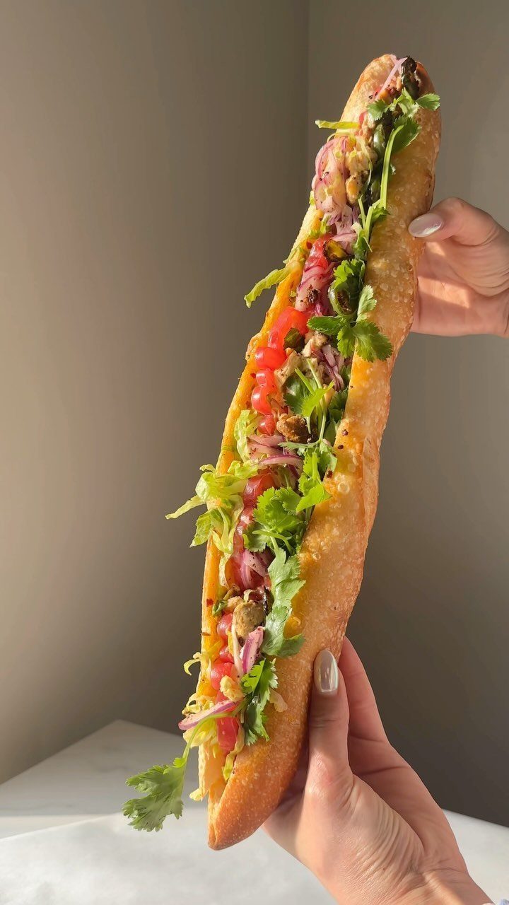 Long baguette sandwich filled with meat, vegetables, and herbs held by two hands.