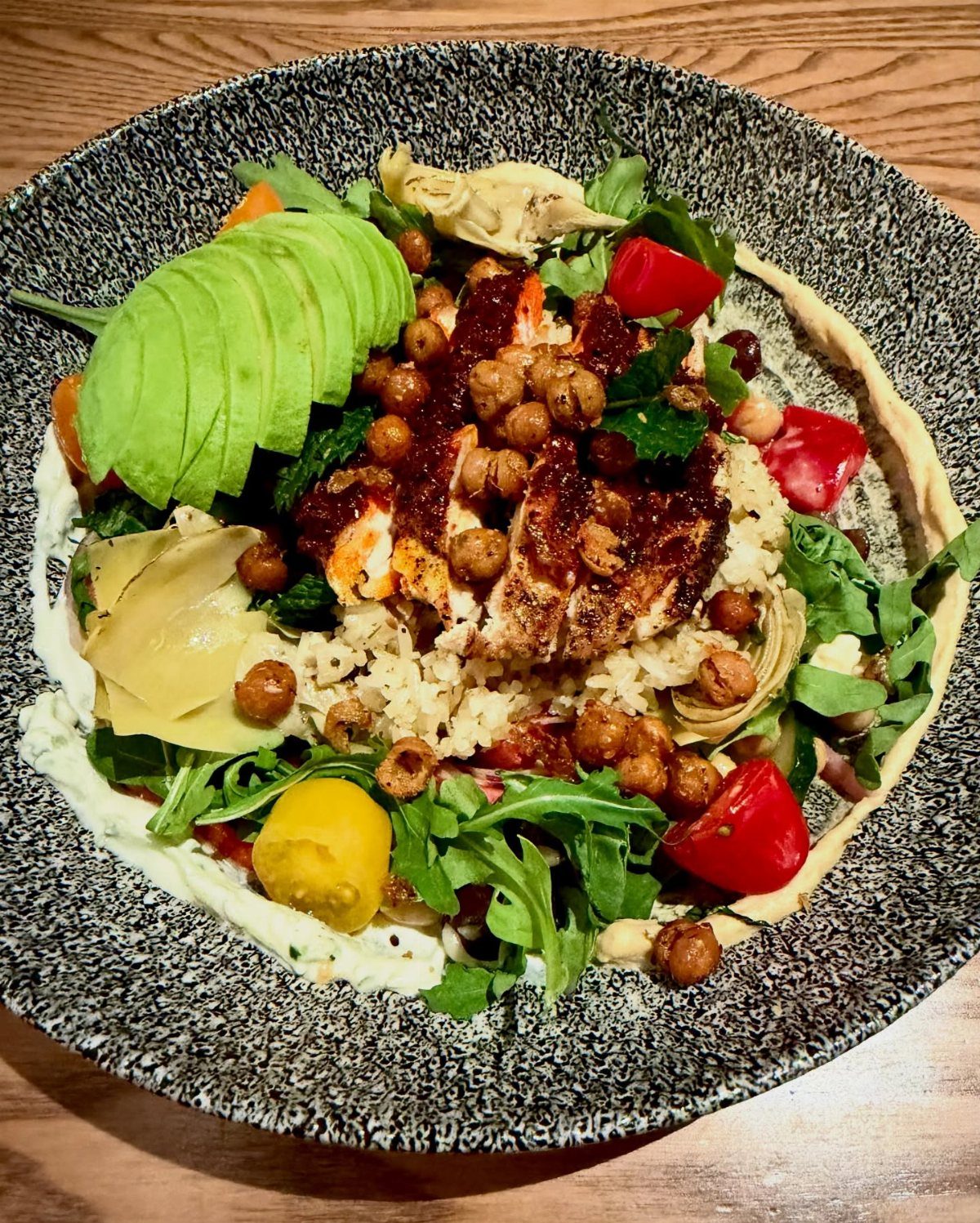 Glazed protein bowl with avocado, chickpeas, cherry tomatoes, and greens in a speckled bowl.