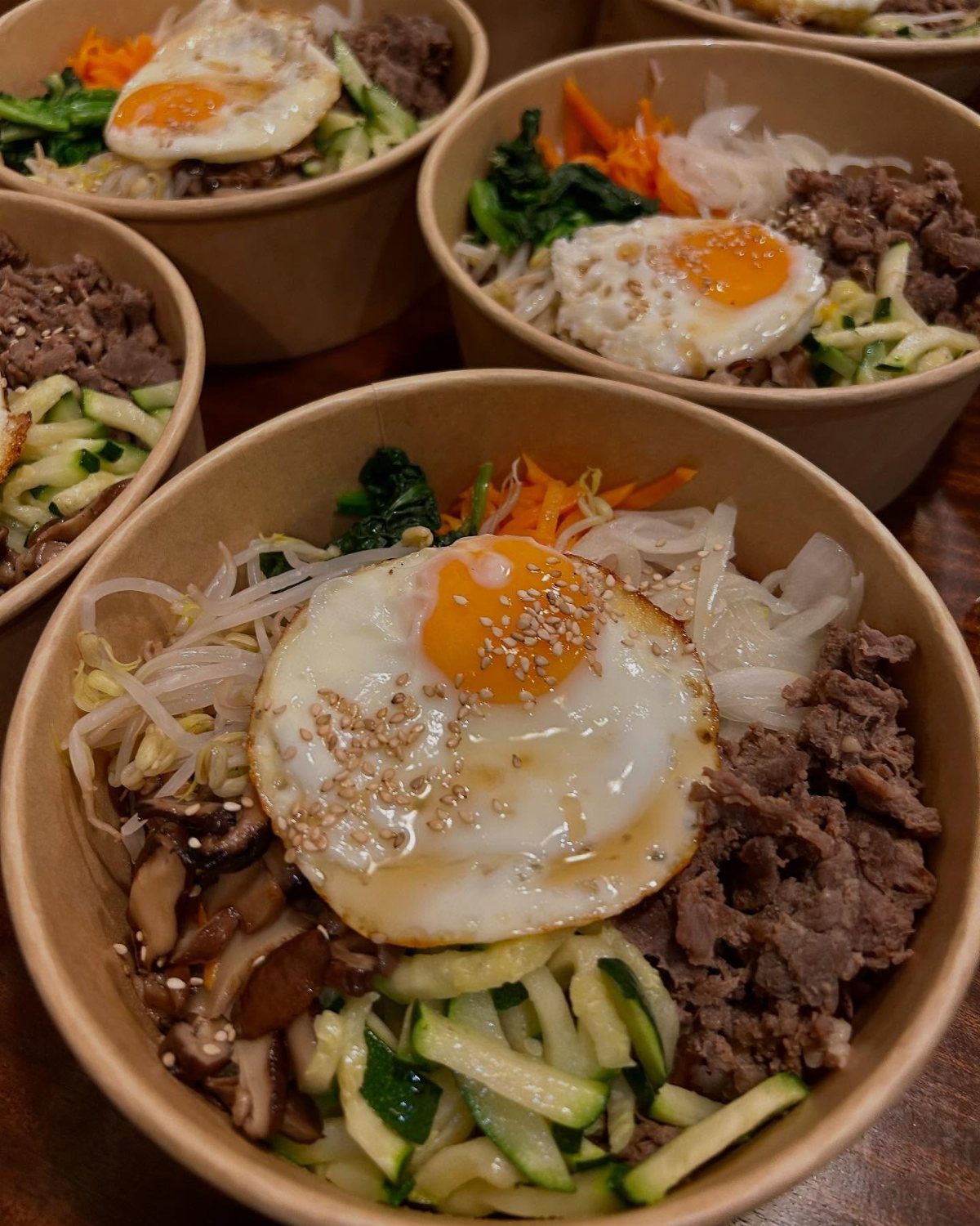 Bibimbap rice bowls with fried eggs, beef, and assorted colorful vegetables in cardboard containers.