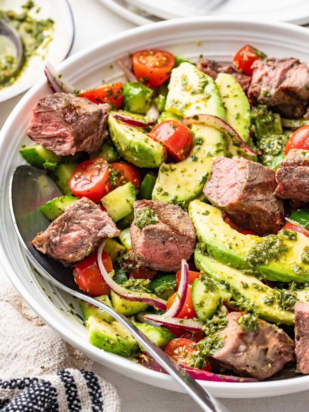 Steak salad with avocado, cherry tomatoes, and pesto in a white bowl.