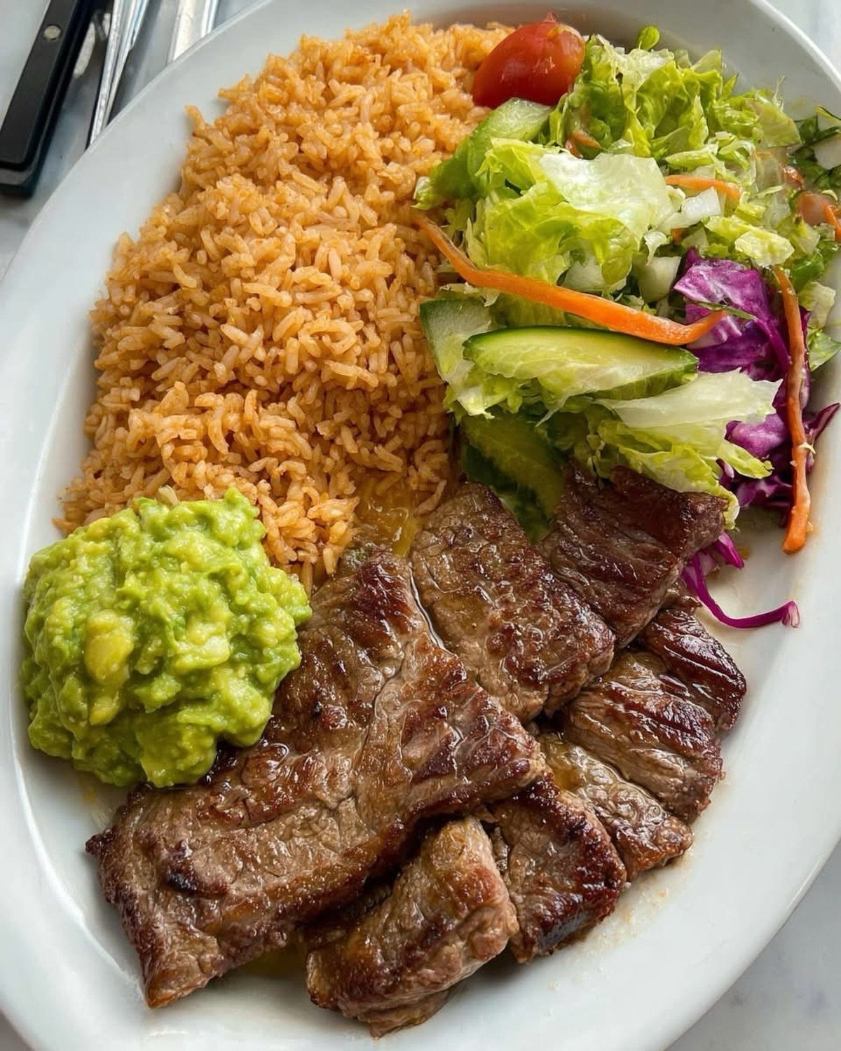 Grilled steak slices with orange rice, guacamole, and a fresh garden salad on a plate.