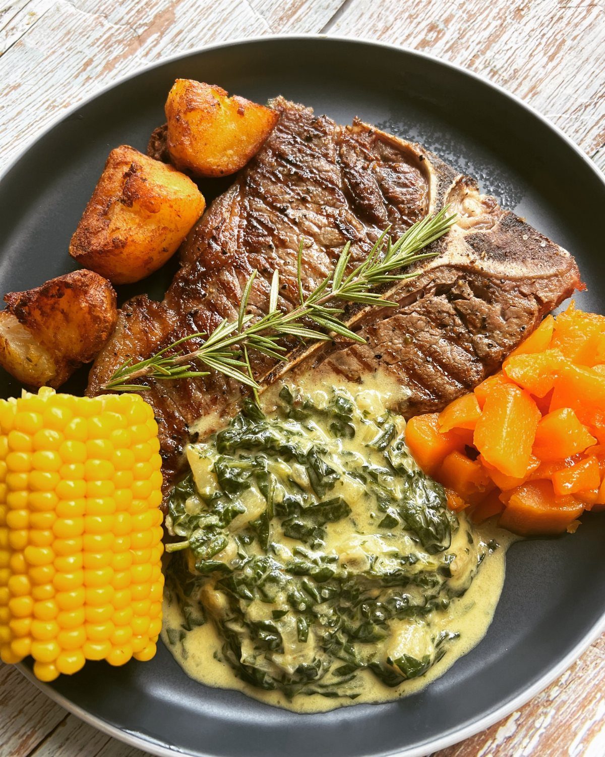 Grilled steak with creamed spinach, roasted potatoes, corn on the cob, and diced vegetables.