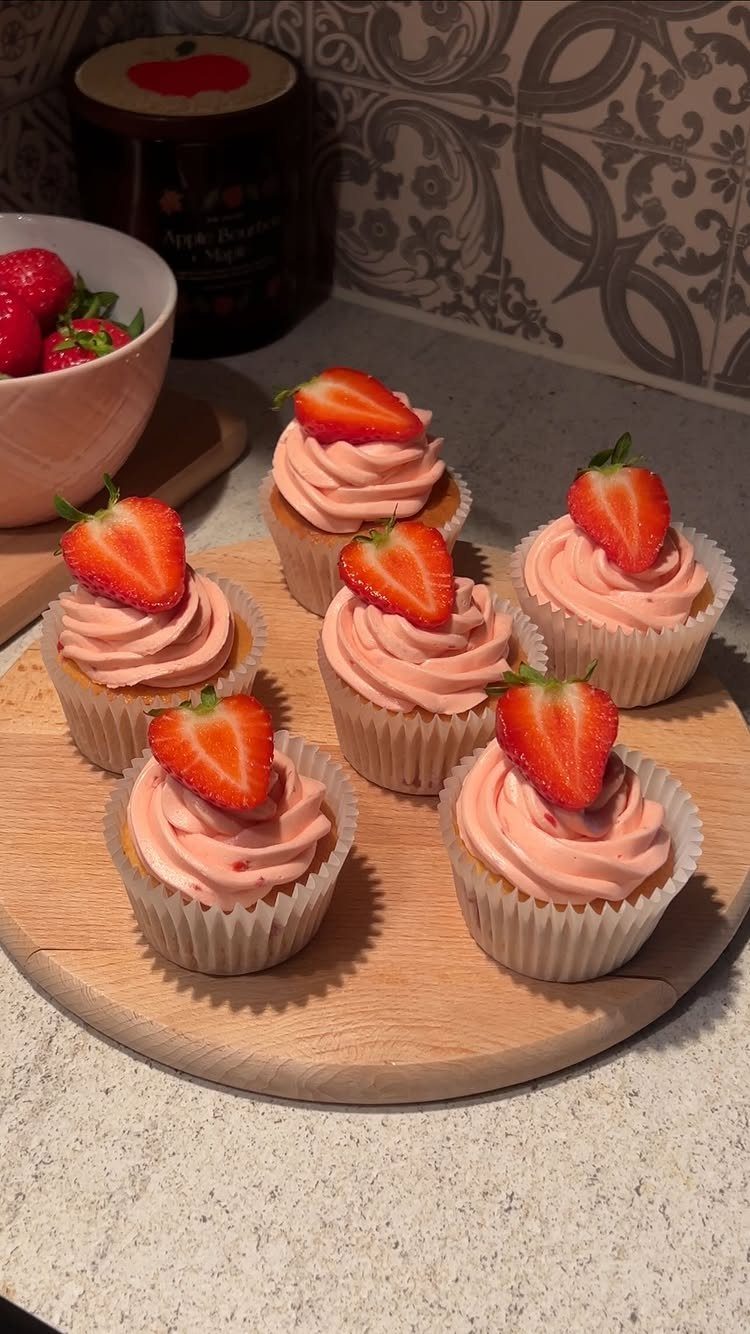 Six pink frosted cupcakes topped with strawberry slices on a wooden serving board.