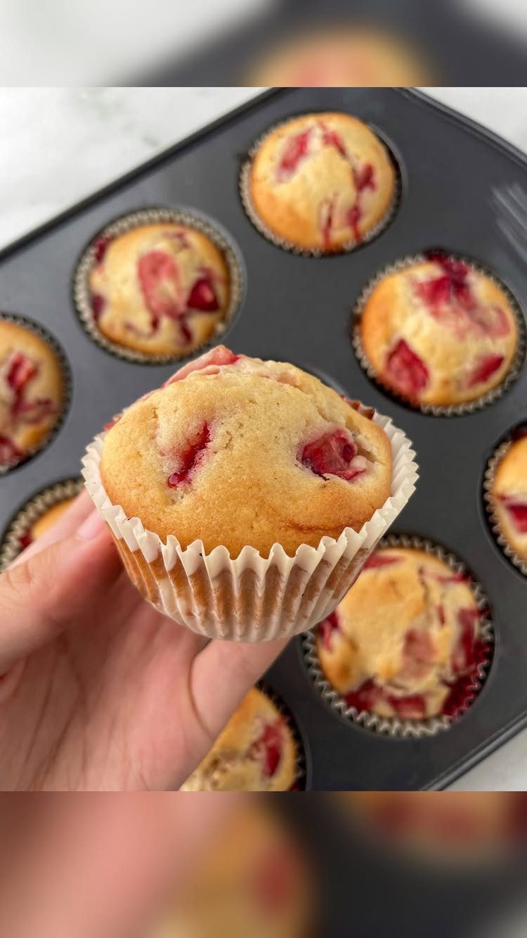 Strawberry muffin held in hand with more muffins in a baking tin.