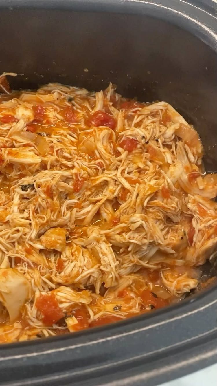 Shredded chicken in a savory red sauce inside a black slow cooker.