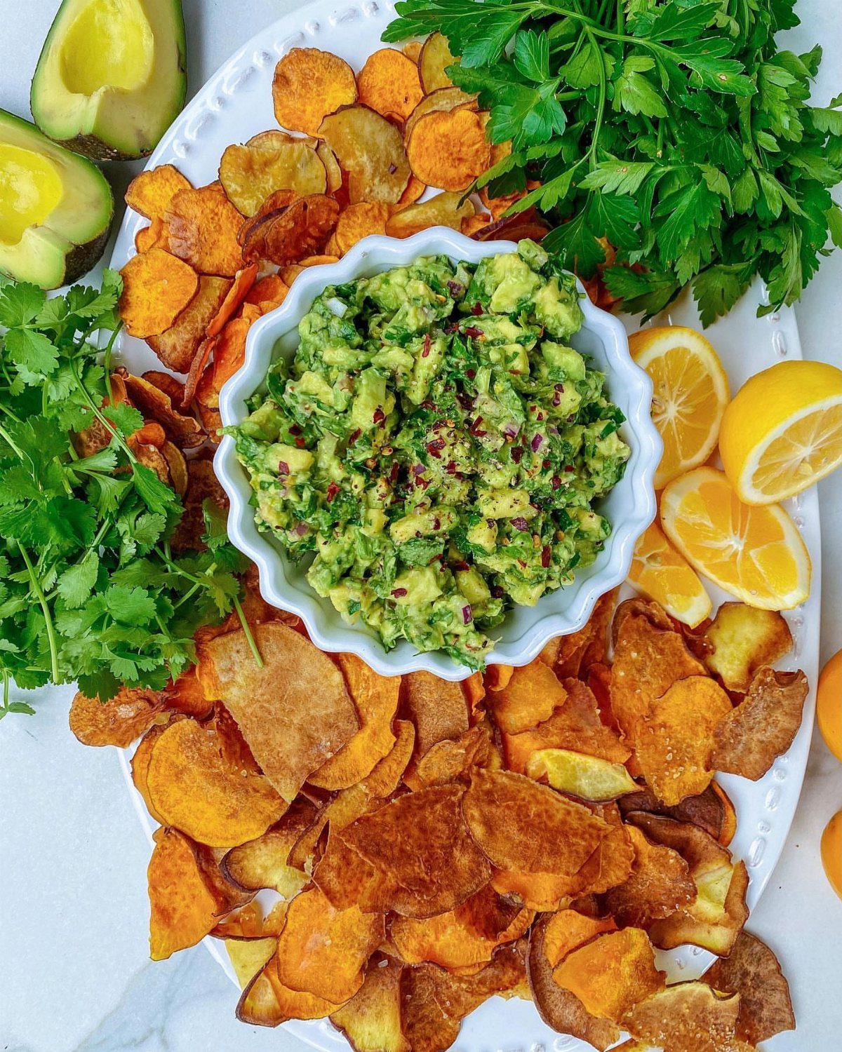 Guacamole bowl surrounded by sweet potato chips, fresh herbs, and lemon slices on a platter.