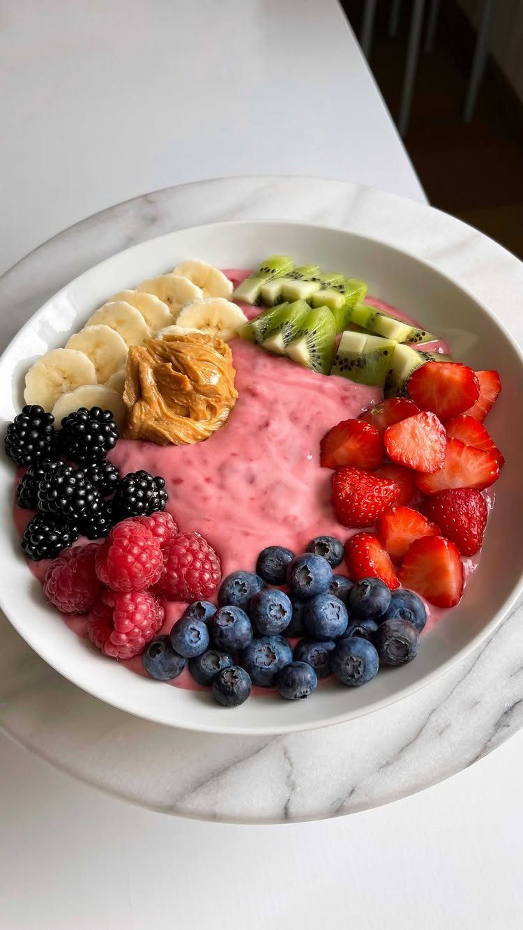 Pink yogurt bowl topped with assorted fresh berries, sliced banana, kiwi, and peanut butter.