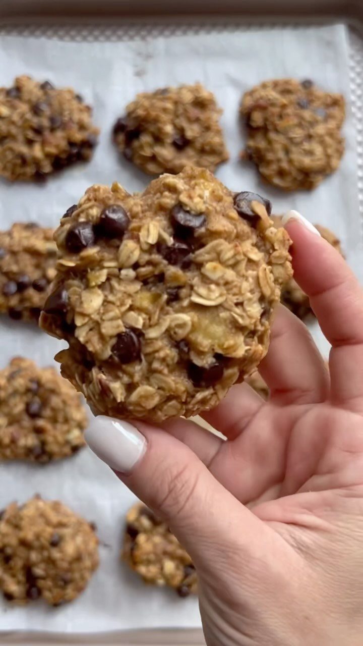 Hand holding a chunky oatmeal cookie with chocolate chips on a baking sheet.