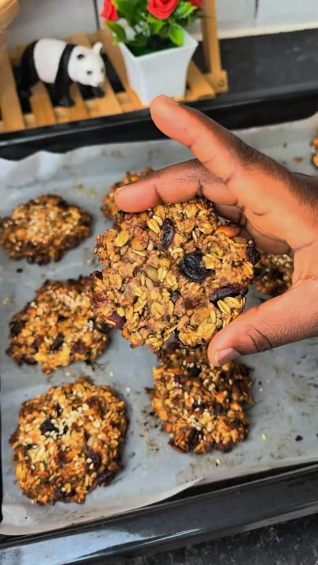 Hand holding a healthy oatmeal cookie with visible raisins and seeds on a tray.