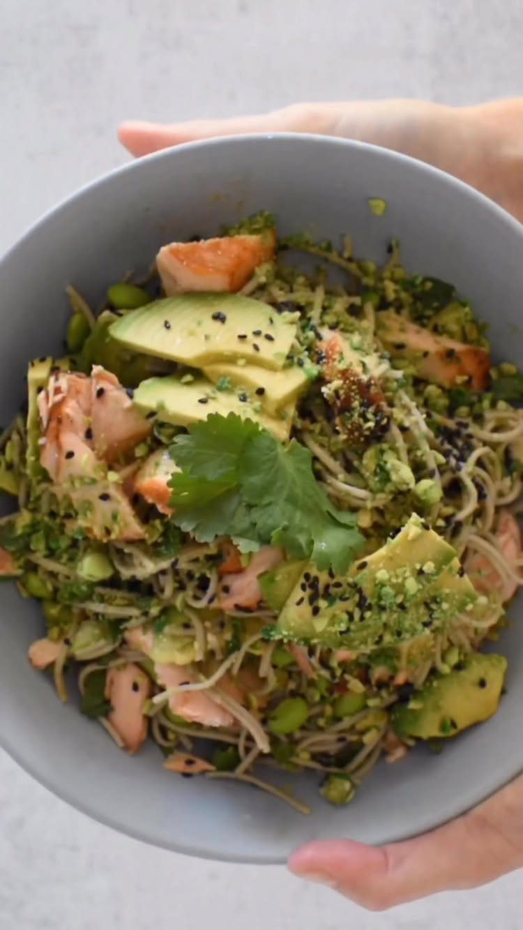 Soba noodle bowl with salmon, avocado, edamame, and cilantro held in two hands.