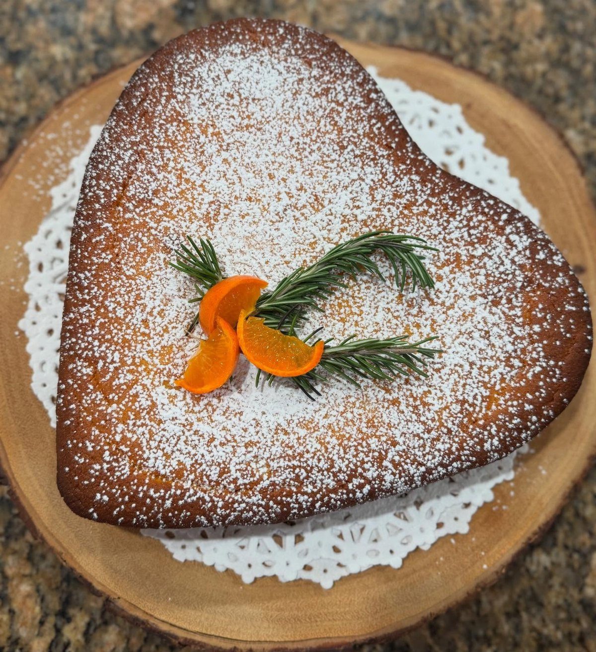 Heart-shaped clementine cake dusted with powdered sugar and garnished with citrus slices and rosemary.