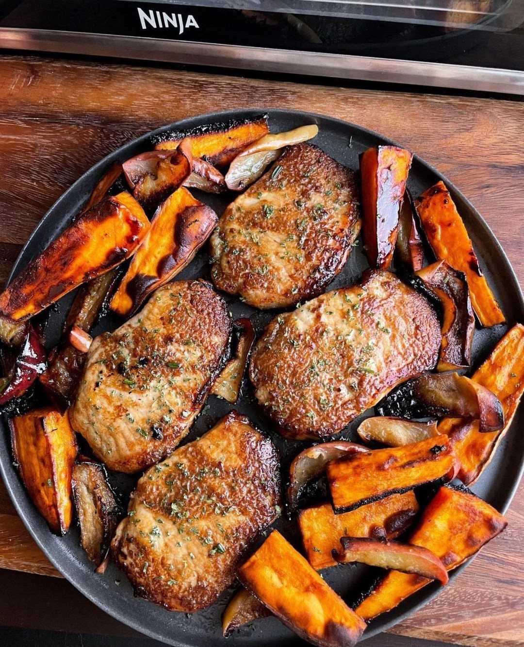 Seasoned pork chops and roasted sweet potato wedges on a dark baking pan.