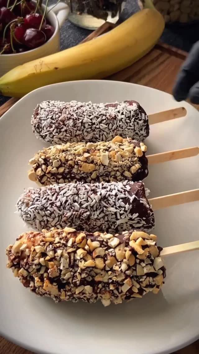 Chocolate covered frozen banana pops with coconut and nuts on a white plate.